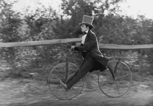 Buster Keaton (wearing a top hat and suit) rides off on a bicycle with no pedals, propelling himself forward by swinging his legs.