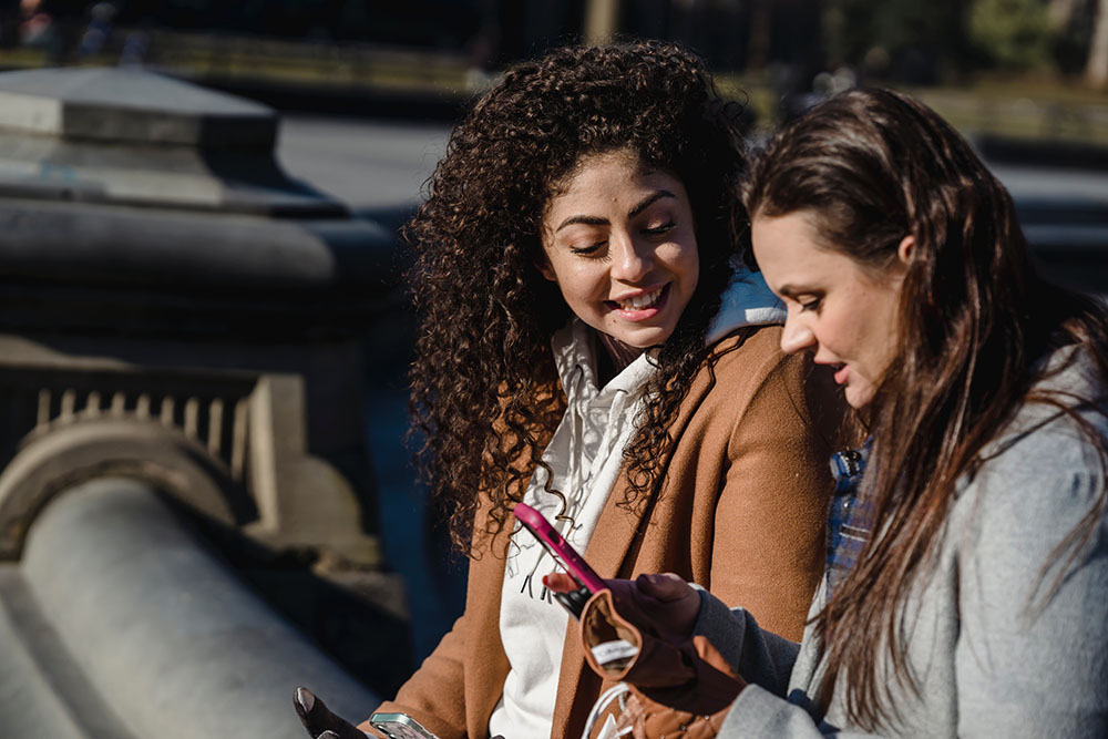 Two young women sitting on a bench. They are looking at one of the womans' phone and smiling
