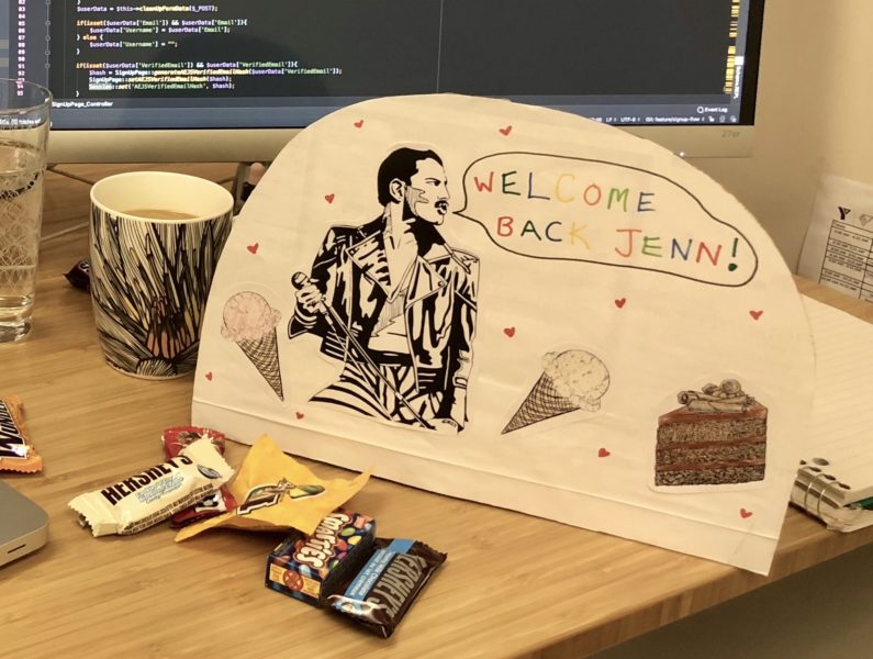 A "welcome back" card sits on a desk infront of a computer and mini chocolate bars. The card has Freddie Mercury, ice cream and cake on it.